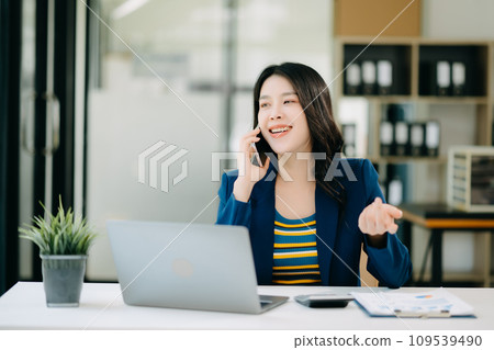 Smiling Asian woman talking on the phone with a customer Young positive female accountant using smartphone talking to team at her desk relaxing with mobile Smiling Asian woman talking on the phone with a customer Young positive female accountant using smartphone talking to team at her desk relaxing with mobile 109539490