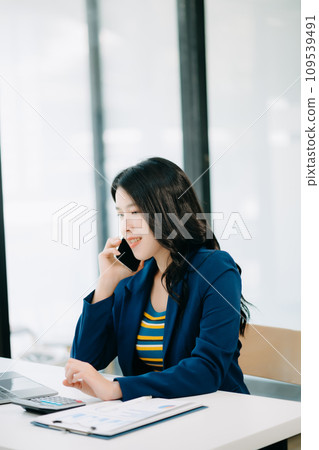 Smiling Asian woman talking on the phone with a customer Young positive female accountant using smartphone talking to team at her desk relaxing with mobile Smiling Asian woman talking on the phone with a customer Young positive female accountant using smartphone talking to team at her desk relaxing with mobile 109539491