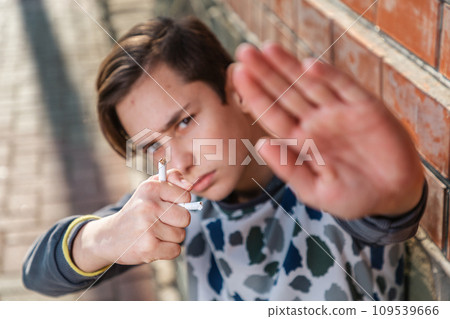 A caucasian teenage boy holds a broken cigarette in his hand and shows a stop sign, protesting against smoking. The concept of adolescent smoking addiction A caucasian teenage boy holds a broken cigarette in his hand and shows a stop sign, protesting against smoking. The concept of adolescent smoking addiction 109539666