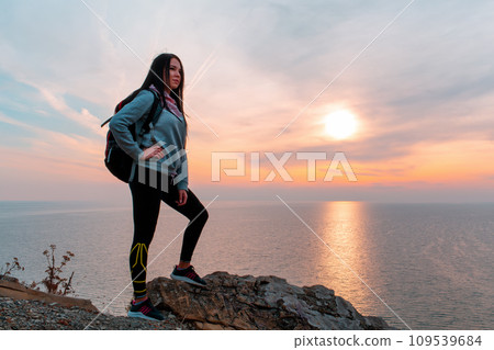 The concept of Hiking and sports recreation. A woman with a backpack on her back poses against the sea and sky. Sunset. Copy space 109539684