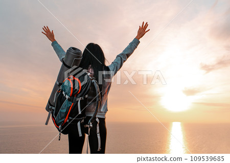 Tourism and sports recreation. The concept of success. A young woman with a tourist backpack behind her back, raises her hands up joyfully. Sunset. Copy space Tourism and sports recreation. The concept of success. A young woman with a tourist backpack behind her back, raises her hands up joyfully. Sunset. Copy space 109539685