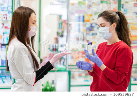 Two young women in medical masks and gloves on the background of a window with pharmacy products, discussing medicine. The concept of virus protection and drug purchase in a pharmacy Two young women in medical masks and gloves on the background of a window with pharmacy products, discussing medicine. The concept of virus protection and drug purchase in a pharmacy 109539687
