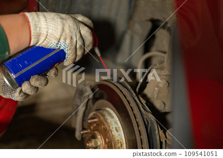 A man is repairing a car in a garage. A man treats rusted bolts with WD-40 solution. Replacing a brake disc on a car in the garage. Concept of a man fixing his car in a garage. 109541051