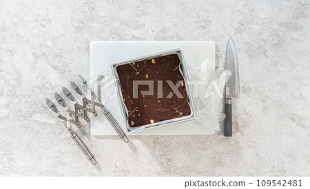 Flat lay. Removing chocolate macadamia fudge from a square cheesecake pan lined with parchment. Flat lay. Removing chocolate macadamia fudge from a square cheesecake pan lined with parchment. 109542481
