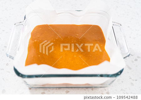 Pouring fudge mixture into the square glass baking pan lined with parchment paper to prepare pumpkin spice fudge. Pouring fudge mixture into the square glass baking pan lined with parchment paper to prepare pumpkin spice fudge. 109542488