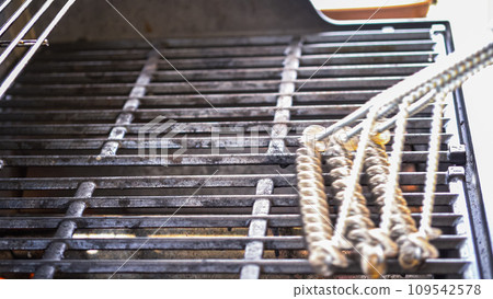 In an outdoor setting, the two-burner gas grill is receiving careful attention before the cooking begins. The grill is meticulously cleaned with a specialized grill brush, ensuring an optimal and 109542578