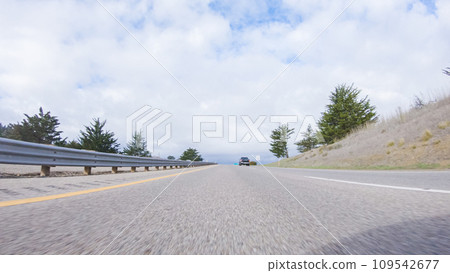 On a crisp winter day, a car cruises along the iconic Highway 101 near San Luis Obispo, California. The surrounding landscape is brownish and subdued, with rolling hills and patches of coastal 109542677