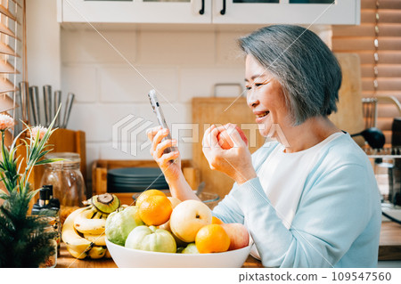 A grandmother, an old woman, is having breakfast in the kitchen. She's smiling, using her smartphone, and eating a red apple. A modern portrait of technology and happiness at home. A grandmother, an old woman, is having breakfast in the kitchen. She's smiling, using her smartphone, and eating a red apple. A modern portrait of technology and happiness at home. 109547560
