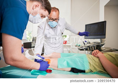 Two doctors making ultrasound diagnostics and injection in knee at hospital. Male specialists looking, examining patient, boy lying on couch. Concept of medicine and healthcare. Two doctors making ultrasound diagnostics and injection in knee at hospital. Male specialists looking, examining patient, boy lying on couch. Concept of medicine and healthcare. 109556470
