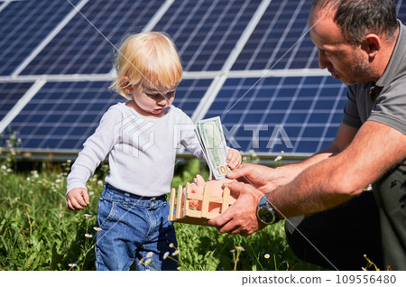 Son and his father putting cash into piggy bank together on background of solar panels. Dad explaining his kid importance of saving money for future. Man and his child investing money to piggy bank 109556480