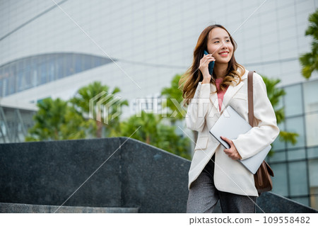 Happy successful Asian businesswoman holding laptop and Talk with cellphone on the Street next to a glass building. successfully work go to against city scene in background of modern office buildings. Happy successful Asian businesswoman holding laptop and Talk with cellphone on the Street next to a glass building. successfully work go to against city scene in background of modern office buildings. 109558482