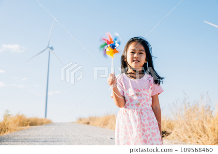 A joyful little girl smiling and running with pinwheels near windmills. Embracing clean energy through playful education in a picturesque wind turbine setting under a beautiful sky. 109568460