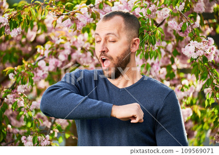 Man allergic suffering from seasonal allergy at spring in blossoming garden at springtime. Bearded young man sneezing and having runny nose in front of blooming tree. Spring allergy concept. Man allergic suffering from seasonal allergy at spring in blossoming garden at springtime. Bearded young man sneezing and having runny nose in front of blooming tree. Spring allergy concept. 109569071