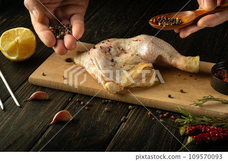 Raw broiler leg on a kitchen cutting board. Adding pepper by hand to a chicken leg before baking at home 109570093