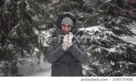 A young woman stands against a background of fir trees under heavy snowfall and rubs her hands against the cold. A girl in glasses and a down jacket with a hood stands and looks at the camera. A young woman stands against a background of fir trees under heavy snowfall and rubs her hands against the cold. A girl in glasses and a down jacket with a hood stands and looks at the camera. 109575406