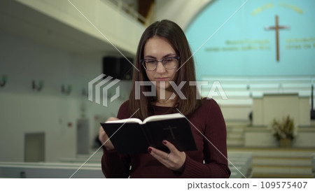 A young woman reads the Bible against the background of a cross in a church. A Protestant girl with glasses reads the Bible in church. A young woman reads the Bible against the background of a cross in a church. A Protestant girl with glasses reads the Bible in church. 109575407