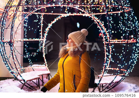 Young woman tourist in knitted hat looks at festive Christmas city decorations. Christmas market, night lights. Young woman tourist in knitted hat looks at festive Christmas city decorations. Christmas market, night lights. 109576416