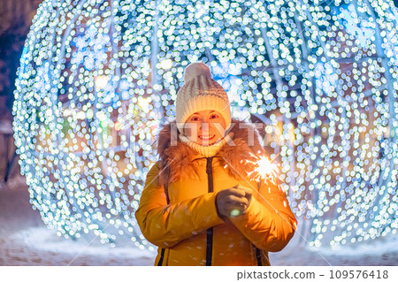 Young woman in knitted hat holds sparkler in her hands. Night festive street, shiny city decorations for Christmas. Young woman in knitted hat holds sparkler in her hands. Night festive street, shiny city decorations for Christmas. 109576418