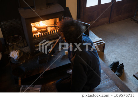 A man putting firewood into a wood-burning stove. An image of preparing for winter in the Japanese winter countryside. A bird's-eye view of a real and polite life. No face. A man putting firewood into a wood-burning stove. An image of preparing for winter in the Japanese winter countryside. A bird's-eye view of a real and polite life. No face. 109576735