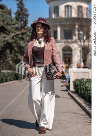 Woman park city. Stylish woman in a hat walks in a park in the city. Dressed in white corset trousers and a pink jacket with a bag in her hands. Woman park city. Stylish woman in a hat walks in a park in the city. Dressed in white corset trousers and a pink jacket with a bag in her hands. 109587157