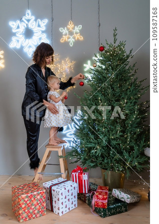 A mother with a 2-year-old daughter decorates the Christmas tree. Mom in a black suit, a girl in a white dress, her daughter hangs a red ball on the Christmas tree. Merry Christmas and New Year A mother with a 2-year-old daughter decorates the Christmas tree. Mom in a black suit, a girl in a white dress, her daughter hangs a red ball on the Christmas tree. Merry Christmas and New Year 109587168