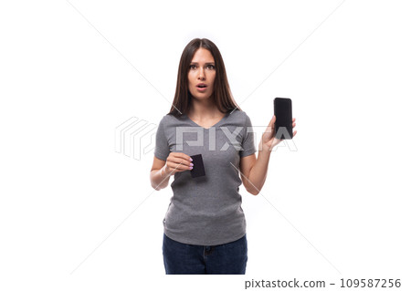 young european brunette woman dressed in a gray basic t-shirt holds a smartphone and a plastic card 109587256