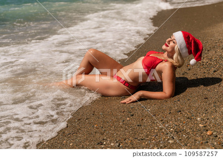 Female beach Santa hat wave coast. beach relaxation seaside. A woman in a red swimsuit enjoying her time on the beach, lying on the sand and being covered by a wave. 109587257