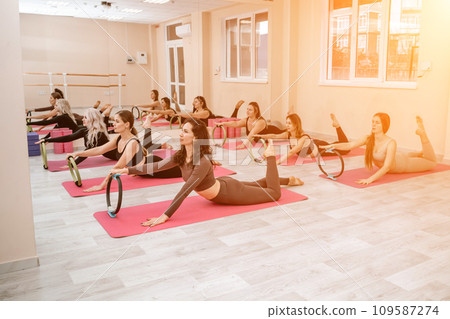 A group of six athletic women doing pilates or yoga on pink mats in front of a window in a beige loft studio interior. Teamwork, good mood and healthy lifestyle concept. A group of six athletic women doing pilates or yoga on pink mats in front of a window in a beige loft studio interior. Teamwork, good mood and healthy lifestyle concept. 109587274