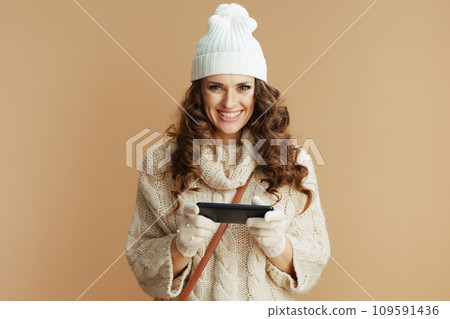 happy woman in beige sweater, mittens and hat using phone app 109591436