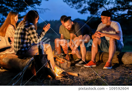 A company of young people sit around a fire at night on the banks of the river and relax in nature. Cooking food on a fire, outdoors 109592604
