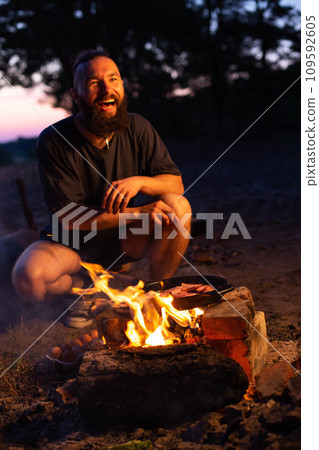 A young man with a beard prepares a dish of bacon with scrambled eggs on a fire. Cooking and outdoor recreation in summer. 109592605