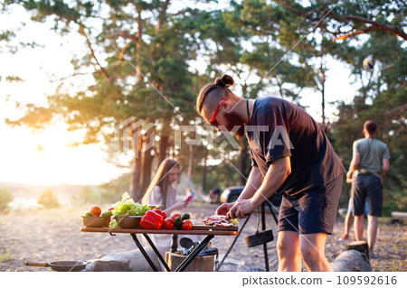 A company of young people prepares a vegetable salad in nature against the backdrop of an evening sunset. Camping, tourism, adventure. Copy space for text 109592616