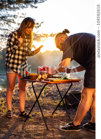 A brutal caucasian man with a beard and black glasses is preparing a vegetable salad in nature against the backdrop of an evening sunset. The girl shoots on the phone. Cooking in nature, tourism A brutal caucasian man with a beard and black glasses is preparing a vegetable salad in nature against the backdrop of an evening sunset. The girl shoots on the phone. Cooking in nature, tourism 109592618