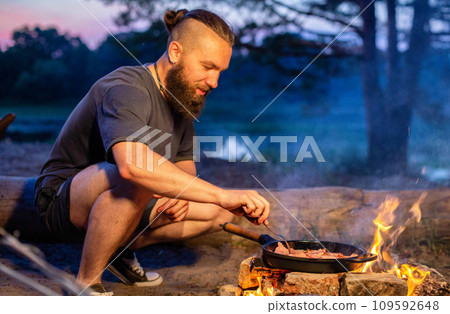 A young man with a beard prepares a dish of bacon with scrambled eggs on a fire. Cooking and outdoor recreation in summer. 109592648