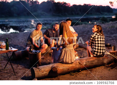 A company of young people sit around a fire at night on the banks of the river and relax in nature. Cooking food on a fire, outdoors A company of young people sit around a fire at night on the banks of the river and relax in nature. Cooking food on a fire, outdoors 109592708