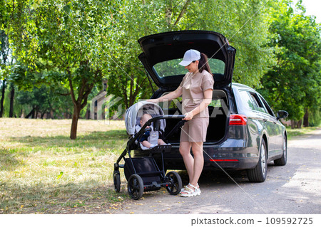 A beautiful girl puts a baby stroller into the open trunk of a car. Family car concept. Sunny weather in summer. 109592725