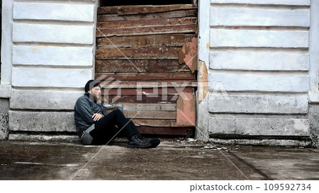 Homeless man sits outside an abandoned building. Refugee needs help. Below poverty line. Illegal immigrant. Tramp wearing in dirty clothes and hat cap. Fugitive. Consequences of drunkenness alcoholism Homeless man sits outside an abandoned building. Refugee needs help. Below poverty line. Illegal immigrant. Tramp wearing in dirty clothes and hat cap. Fugitive. Consequences of drunkenness alcoholism 109592734