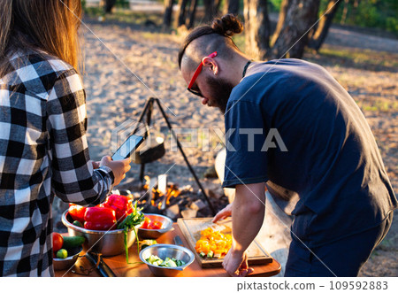 Stylish man with a beard and black glasses cuts fresh vegetables peppers and tomatoes to prepare a salad in nature. Vacation with friends in nature. Tourism as a hobby. Evening sunset. 109592883