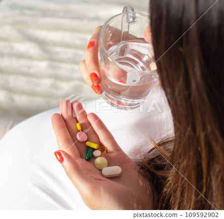 A pregnant girl holds a handful of pills and a glass of water in her hand. The concept of taking vitamins and minerals during pregnancy. Biologically active food supplement and folic acid. 109592902