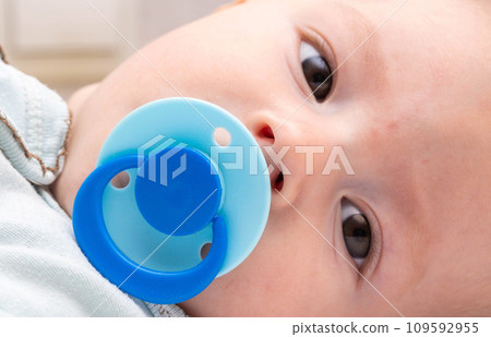 A 5 month old baby with a blue pacifier in his mouth looks at the camera. The concept of the habit of sucking on the nipple, the sucking reflex in babies. A 5 month old baby with a blue pacifier in his mouth looks at the camera. The concept of the habit of sucking on the nipple, the sucking reflex in babies. 109592955