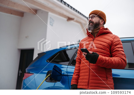 Man charging electric car during cold snowy day, using electric vehicle charging app, checking battery life, energy consumption on smart phone. Charging and driving electric vehicles during winter Man charging electric car during cold snowy day, using electric vehicle charging app, checking battery life, energy consumption on smart phone. Charging and driving electric vehicles during winter 109593174