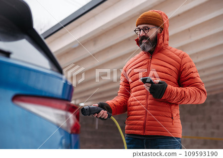 Man charging electric car during cold snowy day, using electric vehicle charging app, checking battery life, energy consumption on smart phone. Charging and driving electric vehicles during winter 109593190