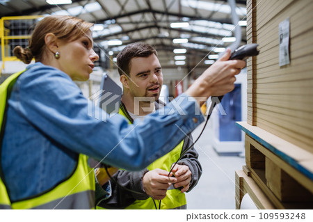 Young man with Down syndrome working in warehouse, female colleague teaching him scan products with scanner. Concept of workers with disabilities, support in workplace. 109593248