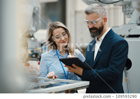Female engineer and male production manager standing in modern industrial factory, talking about production. Manufacturing facility with robotics and automation. Female engineer and male production manager standing in modern industrial factory, talking about production. Manufacturing facility with robotics and automation. 109593290
