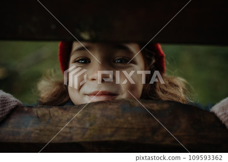 Portrait of little girl hiding behind wooden fence, looking between wooden boards. 109593362