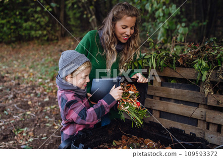 Boy helping mother to put kitchen waste, peel and leftover vegetables scraps into composter in the garden. Concept of composting kitchen biodegradable waste. 109593372