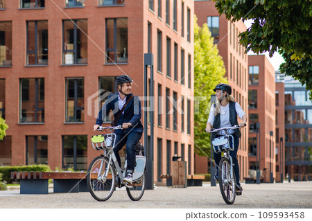 Spouses commuting through the city, riding bike on street. Middle-aged city commuters traveling from work by bike after a long workday. Spouses commuting through the city, riding bike on street. Middle-aged city commuters traveling from work by bike after a long workday. 109593458