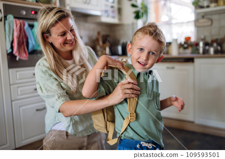 Mother saying goodbye to son before school, helping him to put backpack on back in kitchen. Mother saying goodbye to son before school, helping him to put backpack on back in kitchen. 109593501
