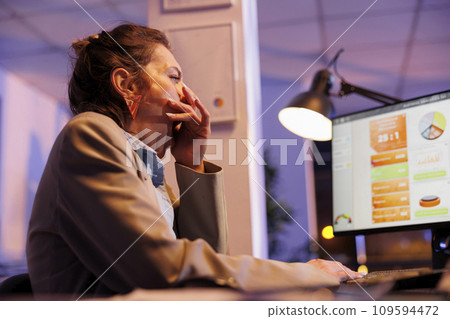 Exhausted manager yawning while analyzing financial graphs report on computer, working overhours at comany investment strategy in startup office. Tired sleepy entrepreneur typing profit progress 109594472