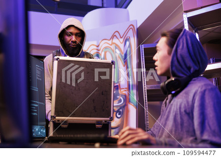 African american hacker opening black box with malicious ransomware program for data breach. Scammer showing case with illegal technology and planning hacking attack with crime partner African american hacker opening black box with malicious ransomware program for data breach. Scammer showing case with illegal technology and planning hacking attack with crime partner 109594477